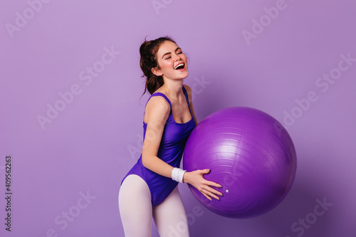 Beautiful girl in purple bodysuit and leggings does exercises with huge fitball on isolated background