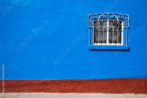 A window covered with an ornate iron cover in a blue wall with brick texture at the bottom.