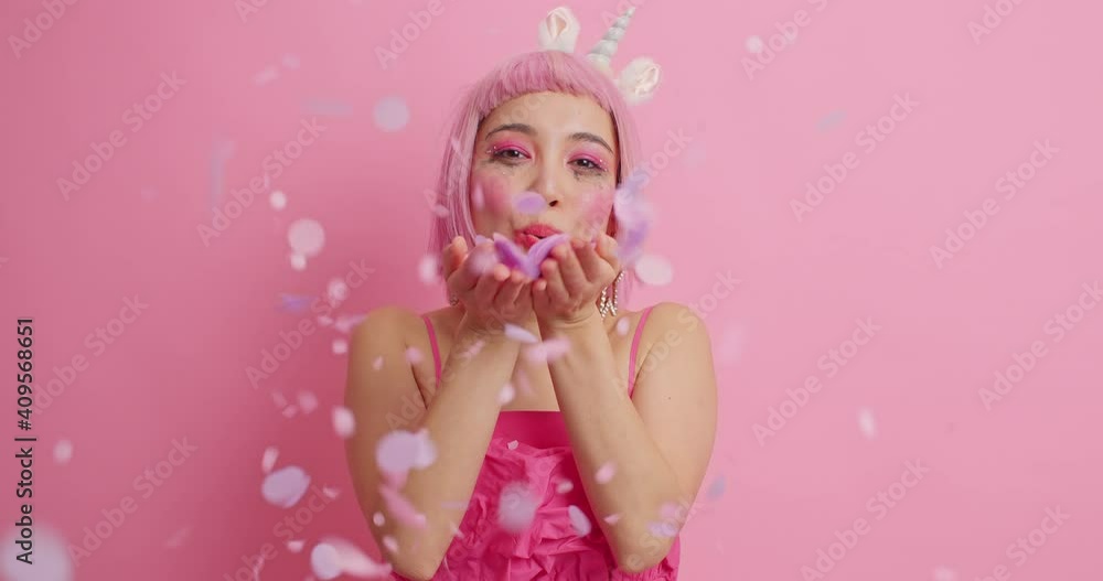 Charming fascinated young Asian woman blows confetti at camera shows her festive mood congratulates friend with birthday wears dress poses against pink studio background. Festive occasion concept Charming fascinated young Asian woman blows confetti at camera shows her festive mood congratulates friend with birthday wears dress poses against pink studio background. Festive occasion concept