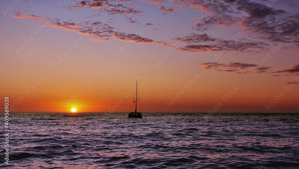 Sunset in the Atlantic Ocean. The sun is low on the horizon. The sky is backlit orange. Beautiful lilac-pink clouds. A lone boat with a mast is visible on the blue surface of the water. South Africa. Sunset in the Atlantic Ocean. The sun is low on the horizon. The sky is backlit orange. Beautiful lilac-pink clouds. A lone boat with a mast is visible on the blue surface of the water. South Africa.