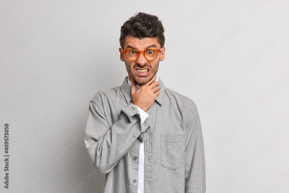 People negative emotions health problems concept. Displeased man keeps hand on throat suffers suffocation clenches teeth from unpleasant feelings wears spectacles and grey shirt poses indoor People negative emotions health problems concept. Displeased man keeps hand on throat suffers suffocation clenches teeth from unpleasant feelings wears spectacles and grey shirt poses indoor