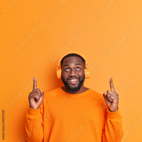 Glad dark skinned bearded male points above with index fingers shows blank space over head listens music via stereo headphones wears casual orange jumper stands indoor alone. Promotion concept