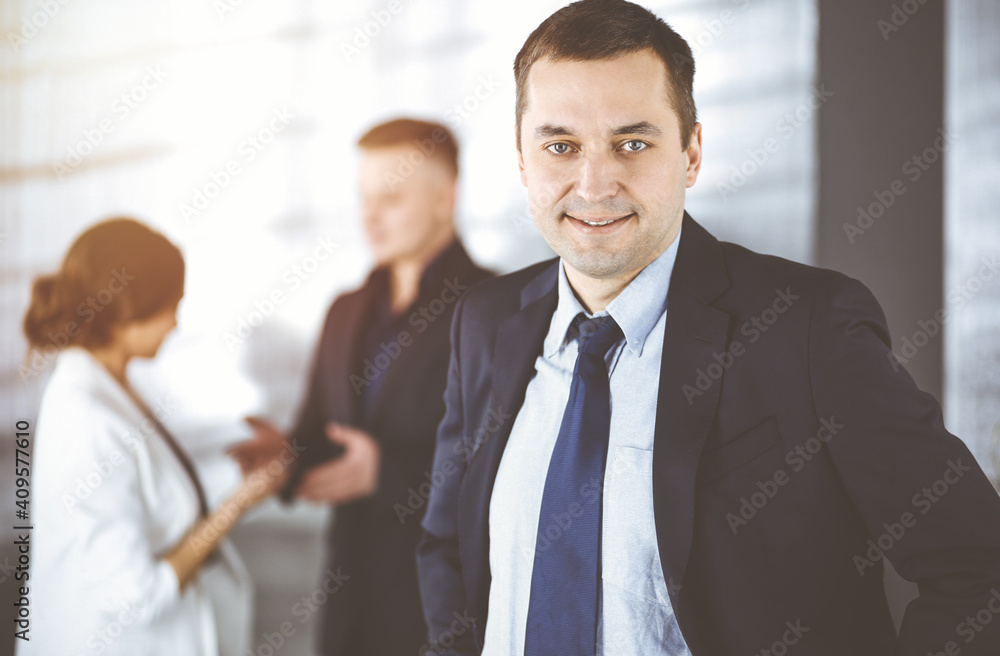 Portrait of a self-confident middle aged businessman in a blue suit, standing in a sunny modern office with his colleagues at the background. Concept of business success Portrait of a self-confident middle aged businessman in a blue suit, standing in a sunny modern office with his colleagues at the background. Concept of business success