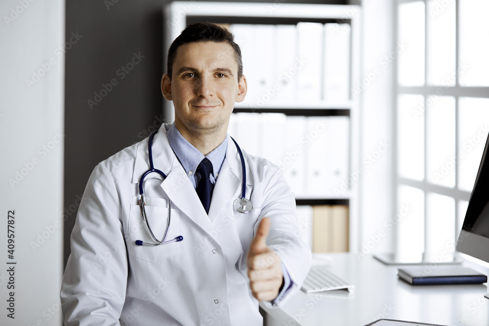 Friendly male doctor sitting and showing OK sign at his working place in clinic. Perfect medical service in hospital. Medicine and healthcare concept
