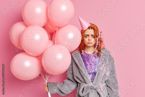 Photo of unhappy young redhead European woman has leaked makeup dressed in soft dressing gown smeared with cream holds bunch of balloons isolated over pink background. Spoiled holiday concept © wayhome.studio 