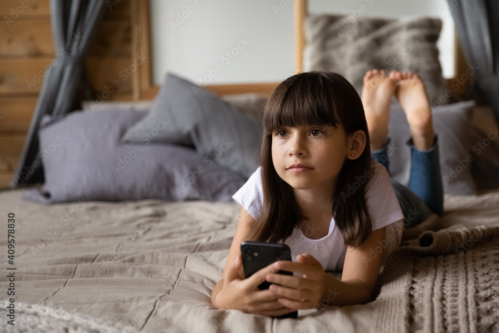 Close up dreamy little girl holding phone, relaxing, lying on cozy bed at home, adorable preschool kid distracted from smartphone, waiting for call or message, enjoying leisure time with gadget Close up dreamy little girl holding phone, relaxing, lying on cozy bed at home, adorable preschool kid distracted from smartphone, waiting for call or message, enjoying leisure time with gadget