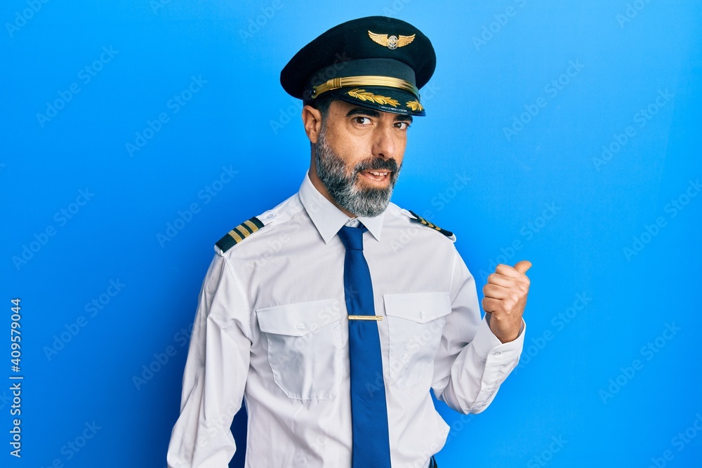 Middle age man with beard and grey hair wearing airplane pilot uniform smiling with happy face looking and pointing to the side with thumb up. Middle age man with beard and grey hair wearing airplane pilot uniform smiling with happy face looking and pointing to the side with thumb up.