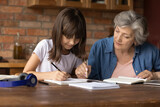 Close up mature grandmother helping to granddaughter with school assignment, focused little girl writing notes, older teacher training pupil, involved in lesson at home, homeschooling concept