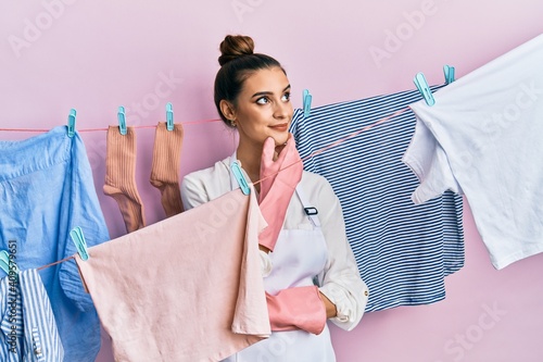 Beautiful brunette young woman washing clothes at clothesline with hand on chin thinking about question, pensive expression. smiling with thoughtful face. doubt concept.