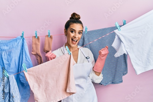 Beautiful brunette young woman washing clothes at clothesline celebrating surprised and amazed for success with arms raised and open eyes. winner concept.