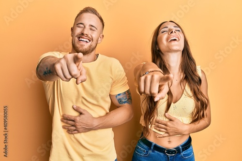 Young couple of girlfriend and boyfriend hugging and standing together laughing at you, pointing finger to the camera with hand over body, shame expression