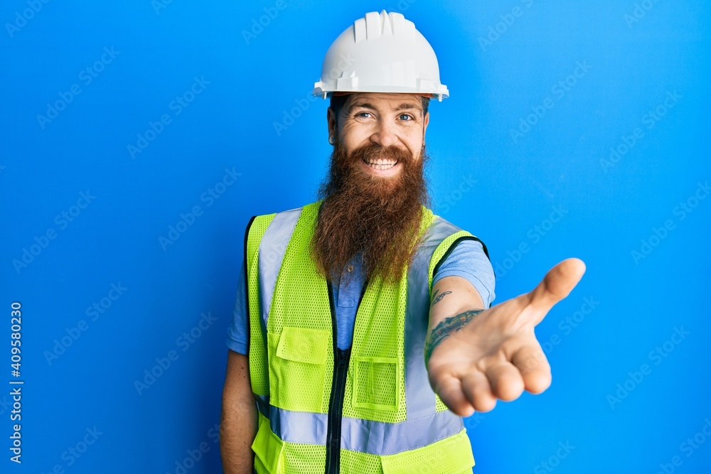 Redhead man with long beard wearing safety helmet and reflective jacket smiling friendly offering handshake as greeting and welcoming. successful business.