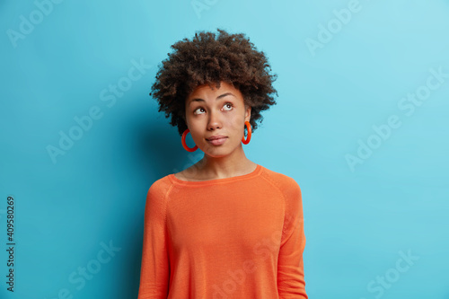 Indoor shot of thoughtful Afro American woman concentrates somewhere pensively has curly hair dreams about something wears casual orange jumper isolated over blue background. Pensive ethnic girl