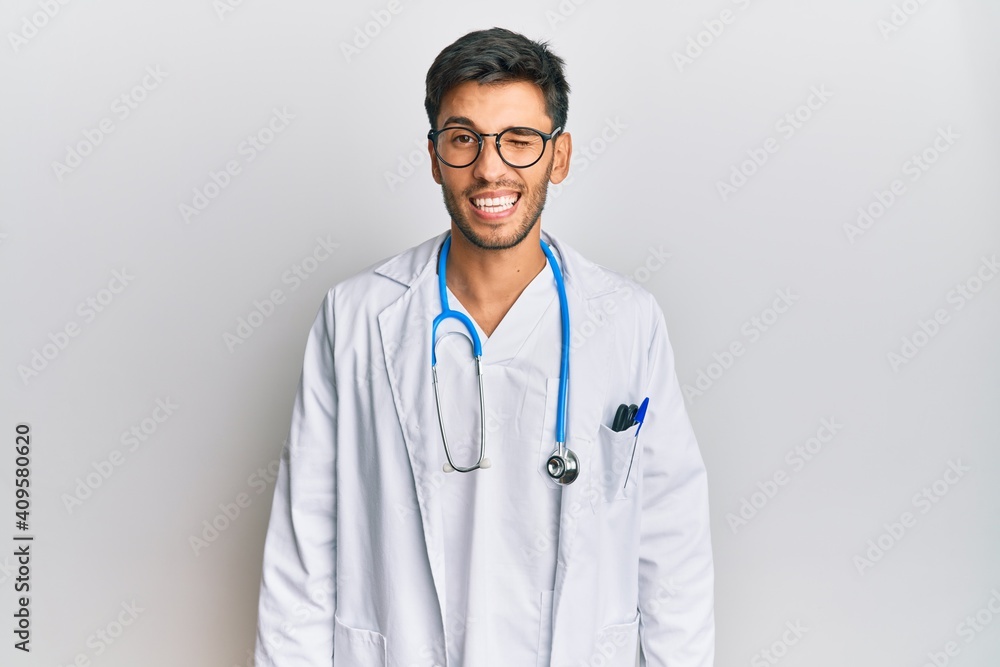 Young handsome man wearing doctor uniform and stethoscope winking looking at the camera with sexy expression, cheerful and happy face. Young handsome man wearing doctor uniform and stethoscope winking looking at the camera with sexy expression, cheerful and happy face.