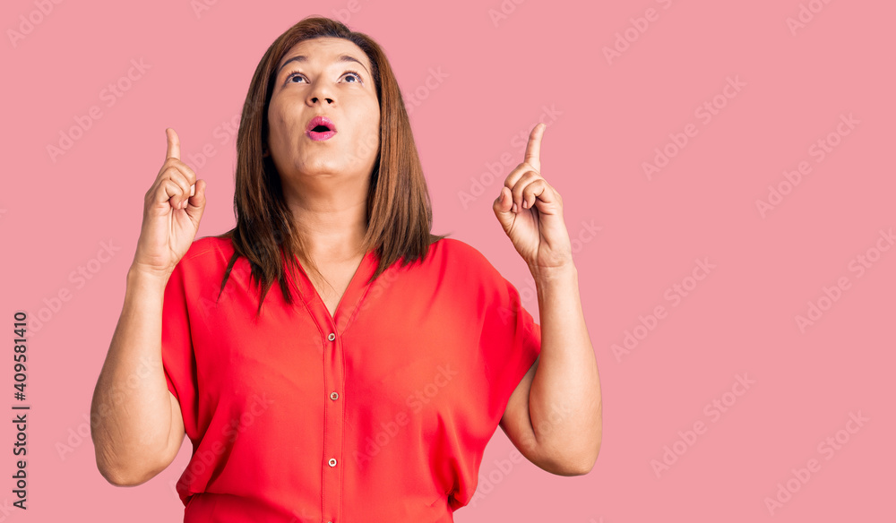 Middle age latin woman wearing casual clothes amazed and surprised looking up and pointing with fingers and raised arms. Middle age latin woman wearing casual clothes amazed and surprised looking up and pointing with fingers and raised arms.