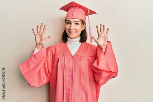 Young caucasian woman wearing graduation cap and ceremony robe showing and pointing up with fingers number ten while smiling confident and happy.