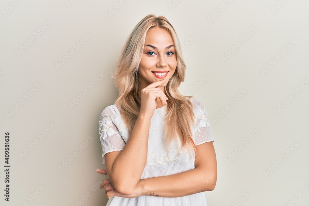 Beautiful caucasian blonde girl wearing white shirt smiling looking confident at the camera with crossed arms and hand on chin. thinking positive. Beautiful caucasian blonde girl wearing white shirt smiling looking confident at the camera with crossed arms and hand on chin. thinking positive.