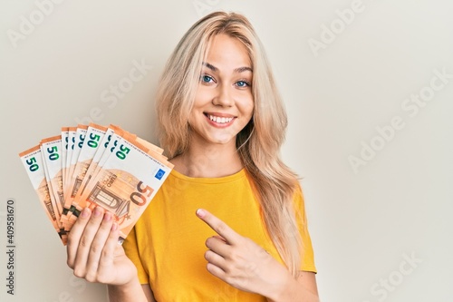 Beautiful caucasian blonde girl holding bunch of 50 euro banknotes smiling happy pointing with hand and finger