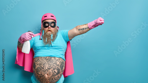 Startled cleaning superhero with detergent going to do washing up stares bugged eyes wears helmet face mask and cape has fat tattoed belly isolated over blue background. Time to tidy up room