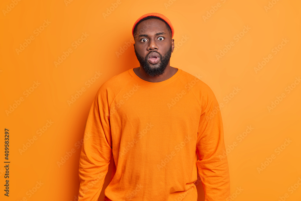 Photo of startled bearded dark skinned man stares at camera with bugged eyes reacts on amazing news sees something horrible afraids of phobia dressed in orange clothes poses indoor. Monochrome Photo of startled bearded dark skinned man stares at camera with bugged eyes reacts on amazing news sees something horrible afraids of phobia dressed in orange clothes poses indoor. Monochrome