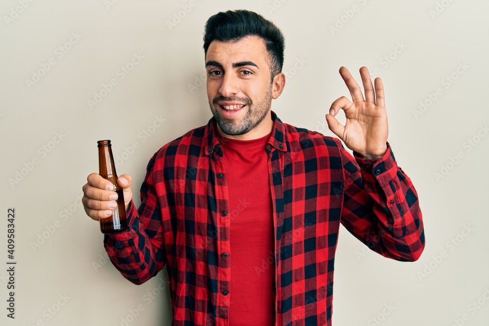 Young hispanic man drinking a bottle of beer doing ok sign with fingers, smiling friendly gesturing excellent symbol Young hispanic man drinking a bottle of beer doing ok sign with fingers, smiling friendly gesturing excellent symbol