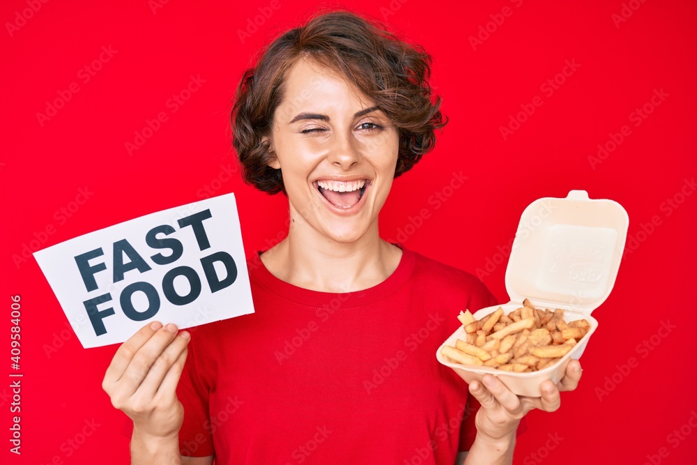 Young hispanic woman holding potato chip and fast food banner winking looking at the camera with sexy expression, cheerful and happy face. Young hispanic woman holding potato chip and fast food banner winking looking at the camera with sexy expression, cheerful and happy face.