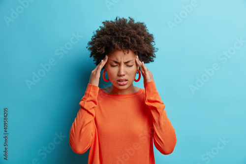 Photo of frustrated young Afro American woman has headache keeps hands on temples suffers unbearable migraine after noisy party wears orange sweater poses indoor isolated on blue background. photo