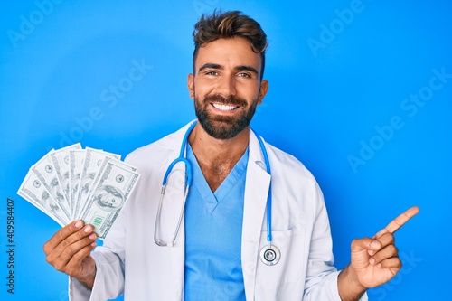 Young hispanic man wearing doctor uniform holding dollars smiling happy pointing with hand and finger to the side photo