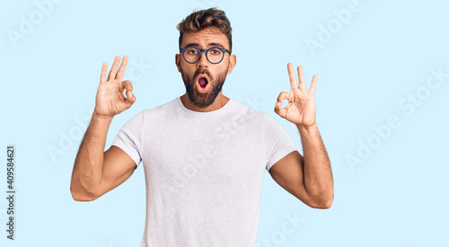 Young hispanic man wearing casual clothes and glasses looking surprised and shocked doing ok approval symbol with fingers. crazy expression
