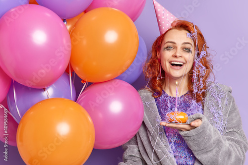 Happy overjoyed redhead woman with leaked makeup enjoys pajama party at home wears birthday cone hat dressing gown smeared with cream smiles broadly holds inflated colorful balloons stands indoor