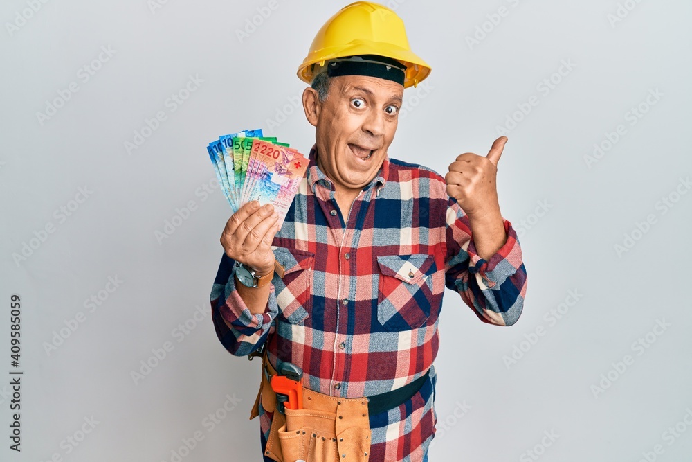 Senior hispanic man wearing handyman uniform and hardhat pointing thumb up to the side smiling happy with open mouth