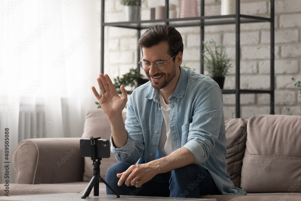 Fototapeta premium Smiling man wearing glasses waving hand, using smartphone on tripod, popular blogger recording vlog, sitting on couch at home, greeting followers, shooting video for channel in social network
