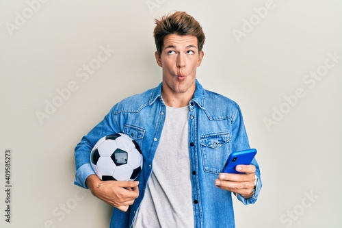 Handsome caucasian man holding football ball looking at smartphone making fish face with mouth and squinting eyes, crazy and comical.
