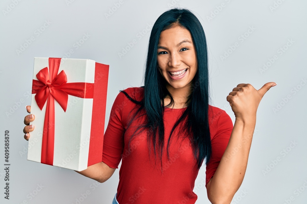 Beautiful hispanic woman holding gift pointing thumb up to the side smiling happy with open mouth Beautiful hispanic woman holding gift pointing thumb up to the side smiling happy with open mouth