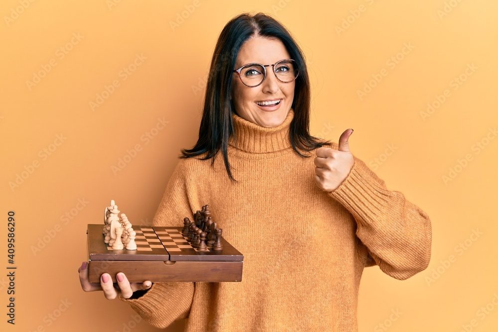 Middle age brunette woman holding chess board smiling happy and positive, thumb up doing excellent and approval sign - obrazy, fototapety, plakaty 
