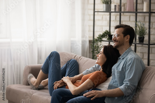 Happy dreamy young couple hugging, relaxing on cozy couch together, renters enjoying leisure time in new apartment, smiling wife and husband planning future, sitting on comfortable sofa at home