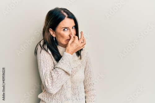 Middle age brunette woman wearing casual clothes smelling something stinky and disgusting, intolerable smell, holding breath with fingers on nose. bad smell