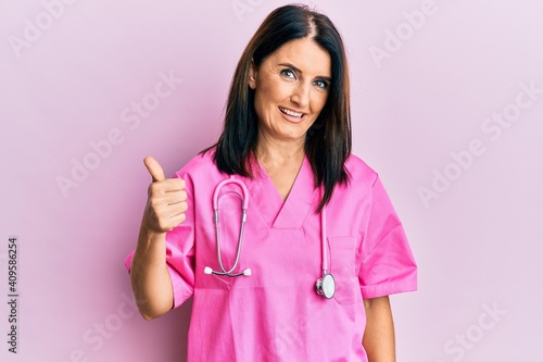 Middle age brunette woman wearing doctor uniform and stethoscope doing happy thumbs up gesture with hand. approving expression looking at the camera showing success.