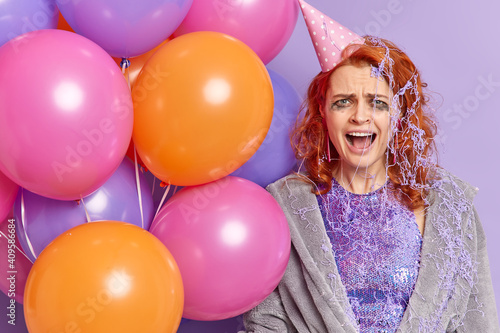 Frustrated redhead woman exclaims with anger frowns face has dissatisfied expression holds multicolored balloons claims about noisy guests on party isolated on purple wall. Celebration concept