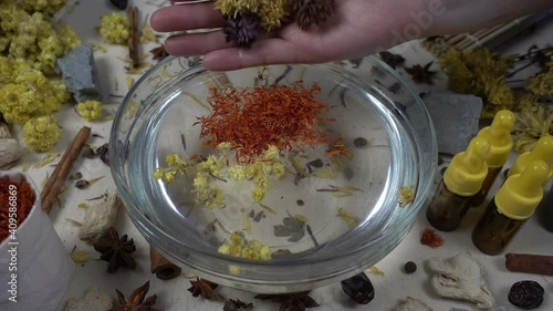 Woman's hand puts aromatic dried flowers and herbs to the glassware with a water. Preparation of natural bath for skin. Alternative medicine, organic cosmetics, aromatherapy and natural care concept.