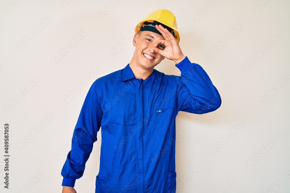 Young hispanic boy wearing worker uniform and hardhat smiling happy doing ok sign with hand on eye looking through fingers