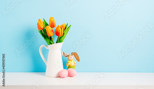 Orange tulip flowers bouquet in jug and pink easter eggs on shelf in front of blue wall. View with copy space