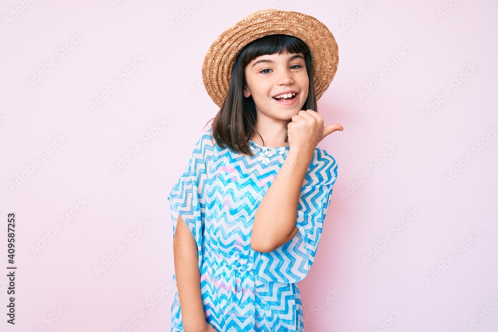 Young little girl with bang wearing summer dress and hat smiling with happy face looking and pointing to the side with thumb up. Young little girl with bang wearing summer dress and hat smiling with happy face looking and pointing to the side with thumb up.