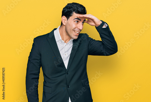 Handsome hispanic man wearing business clothes very happy and smiling looking far away with hand over head. searching concept.