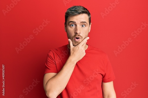Handsome caucasian man wearing casual red tshirt looking fascinated with disbelief, surprise and amazed expression with hands on chin