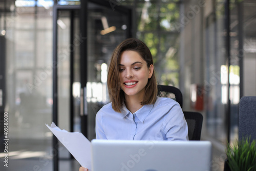 Attractive cheerful business woman checking paper documents in office, working on laptop.