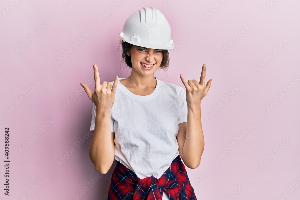 Young caucasian woman wearing hardhat shouting with crazy expression doing rock symbol with hands up. music star. heavy concept. Young caucasian woman wearing hardhat shouting with crazy expression doing rock symbol with hands up. music star. heavy concept.