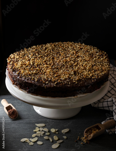 A cocoa cake filled with fruit jam and covered with chopped hazelnuts on a white stand and black background