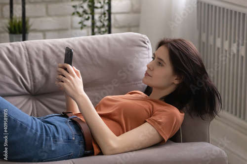 Close up calm young woman lying on cozy couch at home, using smartphone, looking at screen, attractive female relaxing with gadget, enjoying leisure time, chatting online, looking at phone screen