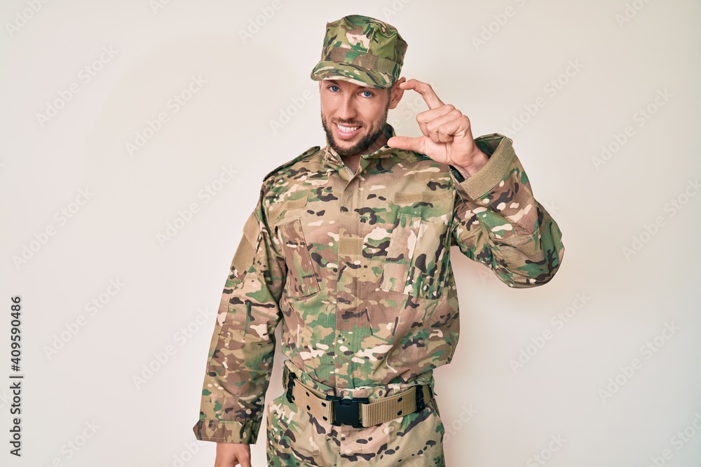 Young caucasian man wearing camouflage army uniform smiling and confident gesturing with hand doing small size sign with fingers looking and the camera. measure concept. Young caucasian man wearing camouflage army uniform smiling and confident gesturing with hand doing small size sign with fingers looking and the camera. measure concept.